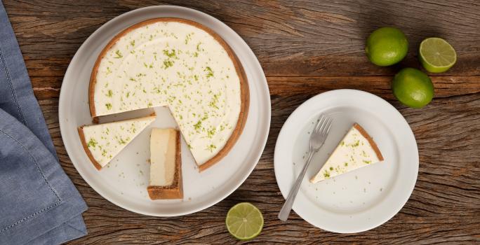 Fotografia em tons de branco e marrom de uma bancada de madeira vista de cima, com um prato branco com uma torta de limão fatiada, ao lado direito prato com uma porção de torta com um garfo e limões, e no esquerdo um guardanapo azul.