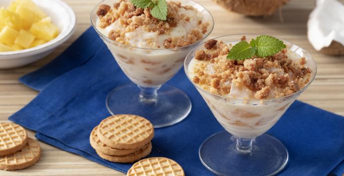 Fotografia em tons de marrom e azul de uma bancada de madeira com um paninho azul, sobre ele duas taças com o creme e biscoitos. Ao fundo um coco e um recipiente redondo com abacaxi.