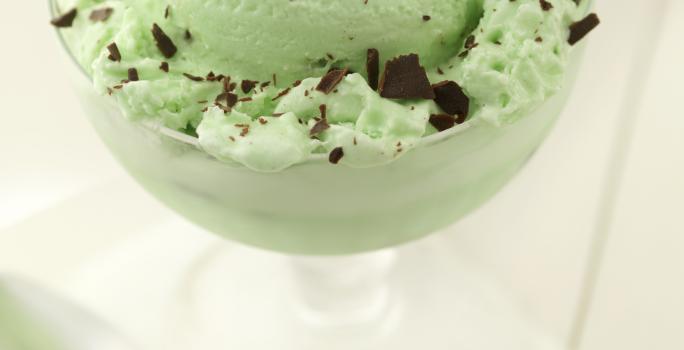 Fotografia de uma bola de sorvete de menta com chocolate em uma taça, que está sobre uma mesa branca. E, ao lado da taça, uma colher de sobremesa.