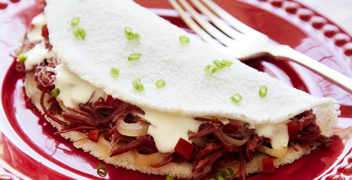Fotografia em tons de branco e vermelho, com prato vermelho ao centro contendo uma tapioca com recheio de carne-seca, queijo de coalho e salpicada de cebolinha, acompanhada de um garfo, sobre guardanapo multicolorido.