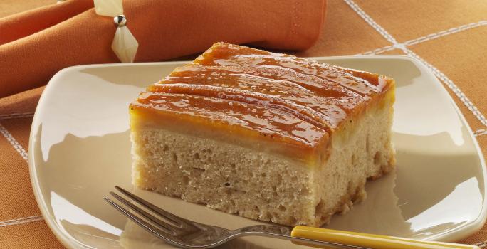 Foto de um prato branco em cima de uma toalha laranja com branco e com um pedaço de bolo de banana dentro com um garfo do lado