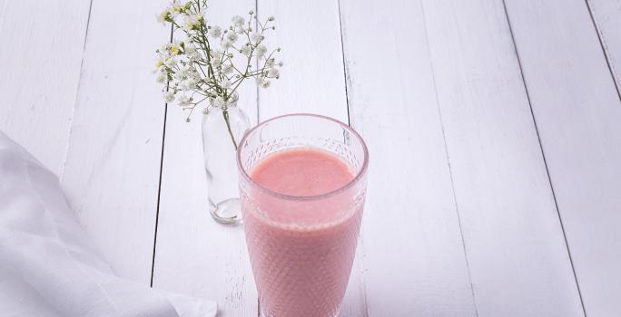 Fotografia em tons de rosa em uma bancada de madeira clara com um copo de vidro ao centro e a vitamina de hortelã com goiaba dentro dele. Ao fundo, um vasinho de vidro com flores brancas pequenas.