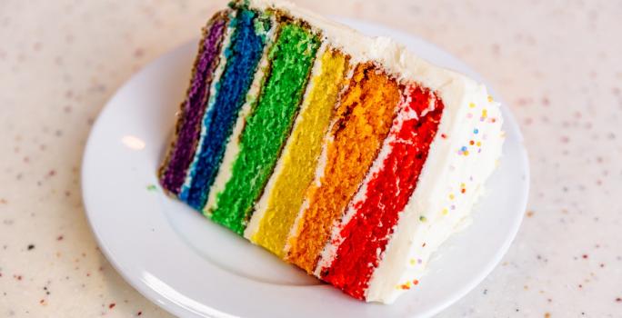 Fotografia de um pedaço de bolo com camadas nas cores vermelho, laranja, amarelo, verde, azul e roxo em cima de um prato branco.