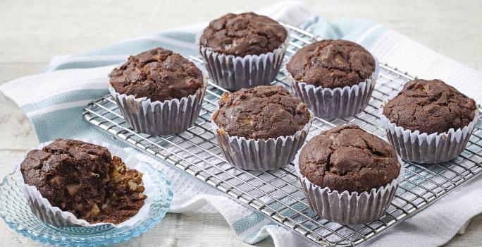 Fotografia em tons de azul em uma mesa de madeira clara com um suporte de ferro com os cupcakes integrais de cacau e banana em forminhas de papel. Ao lado, um pratinho de vidro com um cupcake cortado ao meio.