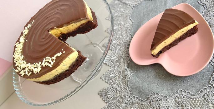 Foto da receita de torta bownie de maracujá, servida em um prato transparente, ao lado de um prato rosa em formato de coração, em uma mesa com uma toalha cinza