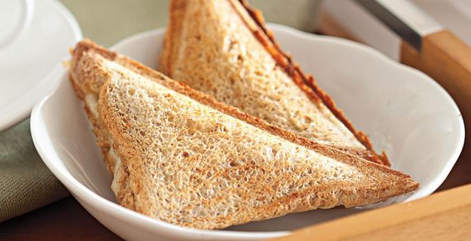 Fotografia em tons de bege em uma mesa com uma toalha bege, um prato redondo fundo branco com dois sanduíches de pão integral tostados e recheados com queijo e lombo.