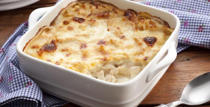 Fotografia em tons de azul em uma bancada de madeira, um pano azul listrado, um recipiente quadrado branco com o frango gratinado dentro dele. Ao lado, uma colher.