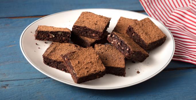 Fotografia em tons de azul, branco e  marrom de uma bancada azul com um prato redondo branco, sobre ele pedaços de brownie de batata-doce. Ao lado um paninho listrado vermelho e branco.