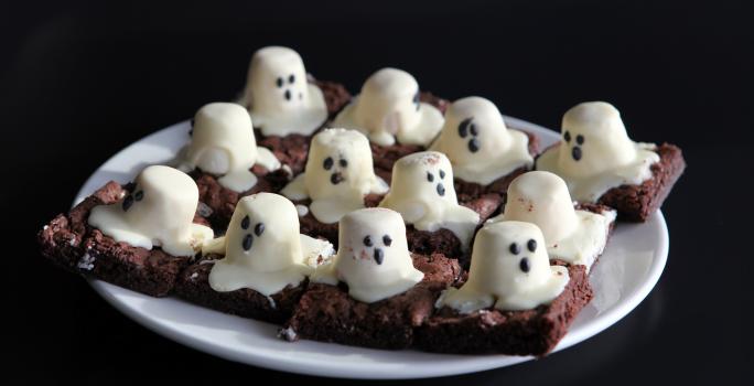 Fotografia de um prato branco com pedaços de brownie de chocolate cortados em quadrados, com marshmallows derretidos em cima representando um fantasma.
