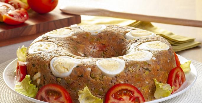 Fotografia em tons de amarelo em uma bancada de madeira de cor branca. Ao centro, um prato branco contendo o cuscuz e ao fundo, há uma tábua de madeira com tomates e uma colher de pau.