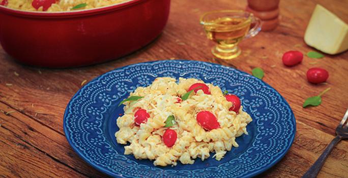 Fotografia em tons de marrom e azul de uma bancada de madeira com um prato azul, sobre ele macarrão com queijo e tomatinhos. Ao lado dois tomatinhos, um pedaço de queijo, um recipiente com azeite e um recipiente redondo vermelho com o macarrão.