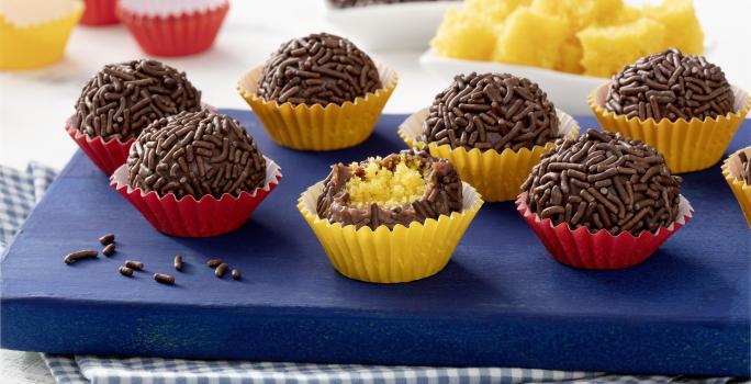 Fotografia em tons de azul, vermelho, amarelo e marrom de uma bancada com um pano quadriculado azul e branco, uma tábua azul, com 7 brigadeiros em cima. Ao centro um brigadeiro mordido, mostrando o recheio de bolo de cenoura.