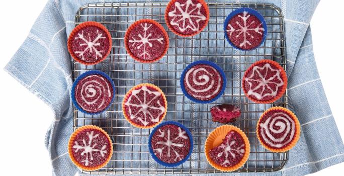 Fotografia em tons de azul e vermelho em um fundo branco com um pano de prato azul jeans com listras brancas, um suporte de gradil com vários cupcakes coloridos de beterraba.