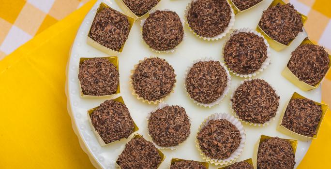 Fotografia em tons de amarelo em uma bancada de madeira com um pano amarelo e outro amarelo xadrez com branco. Em cima deles, um suporte para bolo branco com vários brigadeiros em forminhas amarelas.