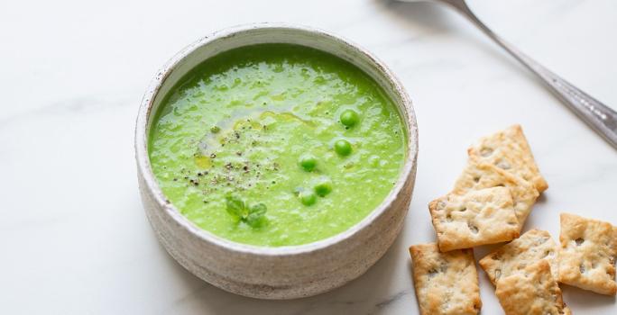 Fotografia em tons de verde em uma bancada de madeira de cor branca. Ao centro, uma tigela contendo a sopa de ervilha com alguns biscoitos de água e sal e uma colher ao lado.