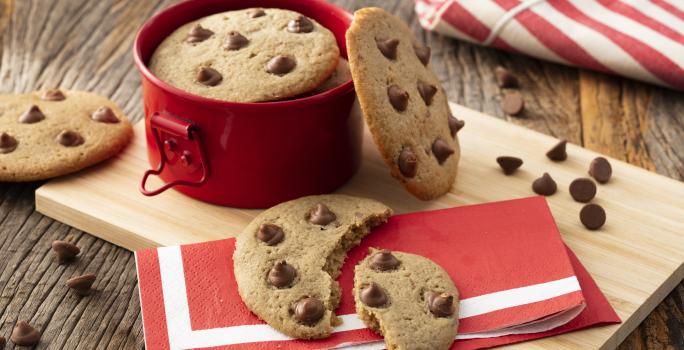 Fotografia em tons de vermelho em uma bancada de madeira com uma tábua de madeira, um guardanapo de papel vermelho com listra branca, um cookie com gotas de chocolate cortado ao meio em cima dele. Ao fundo, uma panelinha vermelha pequena com mais cookies.