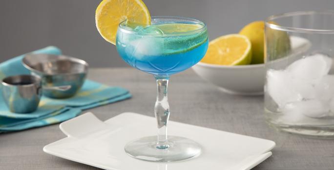 Foto em tons de azul da receita do drink Lagoa Azul servida em uma taça de vidro alta sobre uma base de porcelana branca. Ao fundo há um copo de vidro com cubos de gelo, além de laranjas cortadas e um pano azul para decoração