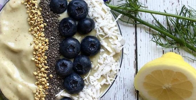 Fotografia em tons de roxo em uma bancada de madeira com um bowl e o creme de avocado dentro dele, decorado com mirtilo, castanha-de-caju e sementes de chia. Ao lado, limão siciliano na mesa.