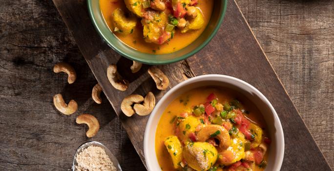Foto de cima de dois pratos redondos e fundos com a receita de moqueca de banana da terra, ambos sobre uma tábua de madeira, com algumas castanhas-de-caju na mesa, uma colher de metal com arroz e um prato com arroz branco