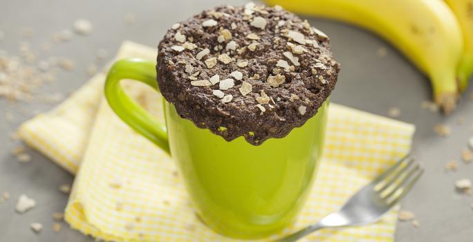 fotografia em tons de cinza, verde e marrom de uma bancada cinza vista de cima. Contém um pano amarelo, uma xicara verde com um bolo de chocolate com cereais por cima e um garfo ao lado e um cacho de bananas