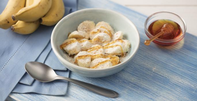 Fotografia em tons de azul em uma mesa de madeira clara com um paninho azul ao lado, um prato branco redondo fundo com as rodelas de banana cobertas com mel, aveia, leite em pó e linhaça. Ao lado, um cacho de bananas e um potinho com mel.