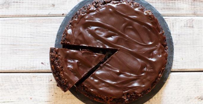 Fotografia em tons de marrom e branco de uma bancada marrom vista de cima, contém um prato redondo e branco com um bolo de chocolate com cobertura de chocolate e uma fatia sendo retirada por uma espátula