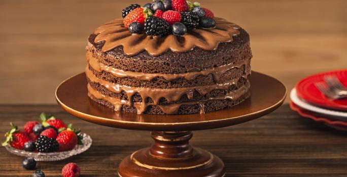 Fotografia em tons de marrom em uma bancada de madeira escura com um suporte para bolo marrom e o naked cake com ganache de cacau e frutas vermelhas em cima. Ao lado, um pratinho de vidro com frutas vermelhas.