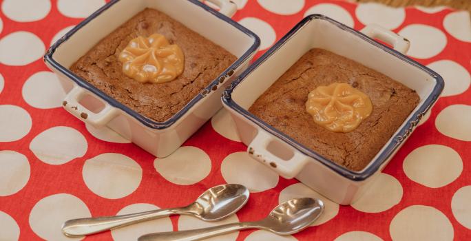 Fotografia em tons de marrom e vermelho de uma bancada de madeira sobre ela um paninho vermelho com bolinhas brancas com duas colheres se sobremesa. Sobre ele dois recipientes quadrados e brancos, dentro o brownie e o MOÇA doce de leite.