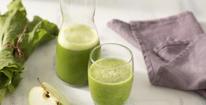Fotografia em tons de verde e roxo de uma bancada branca, sobre ela dois copos de vidro com suco verde. Ao lado um paninho roxo, uma fatia de maçã verde e folhas de couve.