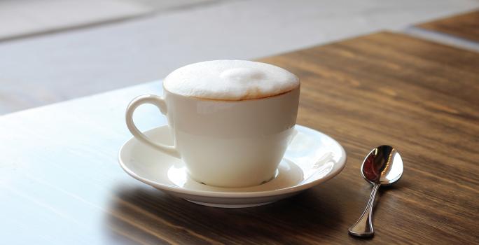 fotografia em tons de marrom e branco tirada de uma bancada marrom. Contém um pratinho branco com uma xicara de café branca, e dentro contém a bebida Cappuccino.