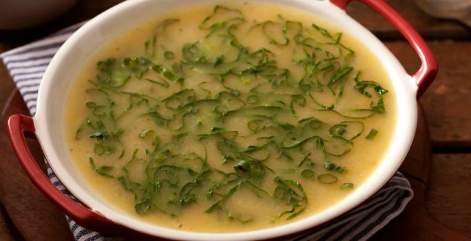 Fotografia em tons de marrom e branco de uma bancada de madeira com paninho listrado azul e branco sobre ele uma panela com sopa de mandioca com couve.