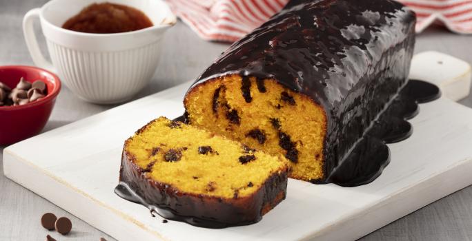 Fotografia em tons de branco e vermelho de uma bancada branca branca com um prato retangular branco, sobre ele um bolo de cenoura. Ao fundo um paninho listrado vermelho e branco e um recipiente redondo branco com calda de chocolate.