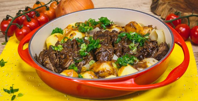 Foto aproximada de uma travessa vermelha com a receita de Carne de Panela, em uma bancada de madeira decorada com cebola, tomate-cereja, um tecido amarelo e algumas cebolinhas verdes