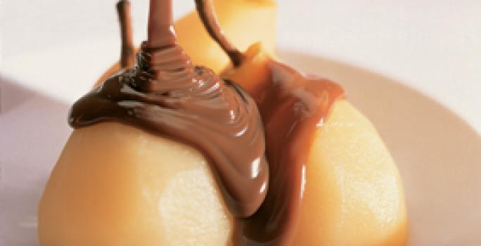 Fotografia em tons de amarelo em uma bancada de madeira branca, um prato redondo raso branco com as peras e calda de chocolate em cima dele. Fotografia em tons de amarelo em uma bancada de madeira branca, um prato redondo raso branco com as peras e calda de chocolate em cima dele.