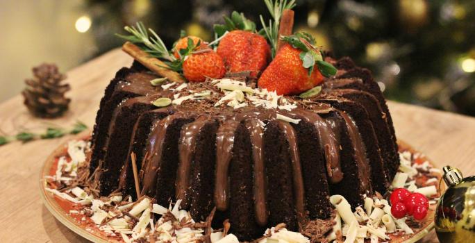 Fotografia em tons de marrom e vermelho de uma bancada de madeira clara, sobre ela um prato vermelho redondo com o bolo de chocolate decorado. Ao lado uma bolinha natalina e uma pinha.