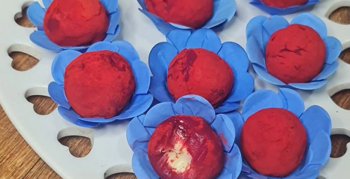Foto em tons de vermelho da receita de brigadeiro red velvet servida em diversas porções sobre embalagens azuis, que se dispõem em cima de uma base em formato de coração branca sobre uma mesa de madeira