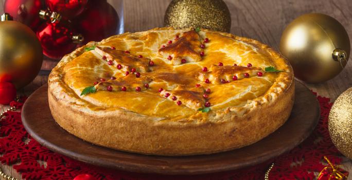 Fotografia em tons de vermelho e dourado em uma bancada de madeira clara, um pano redondo natalino vermelho, um prato de madeira com o empadão em cima dele. Ao lado, bolas e enfeites de Natal.