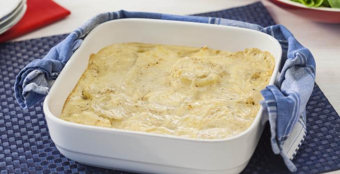 Fotografia em tons de azul e branco de uma bancada vista de cima. Contém um pano azul com um recipiente quadrado branco com alças e dentro as batatas gratinadas. Ao lado um pano azul.