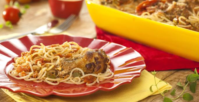 Fotografia em tons de amarelo e vermelho em uma bancada de madeira clara, panos amarelo e vermelho, um recipiente amarelo com o macarrão espaguete com asa de frango dentro dele, um prato vermelho com um pouco do macarrão.