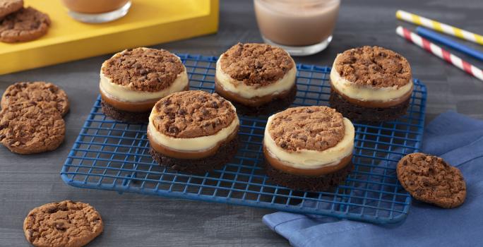 Foto de uma bancada cinza com uma grade contendo cinco doces circulares em camadas diferentes e decorados com cookies Nescau. Além disso há um tecido azul à direita e, ao fundo, canudos coloridos de papel, uma bandeja amarela e copos com leite e Nescau