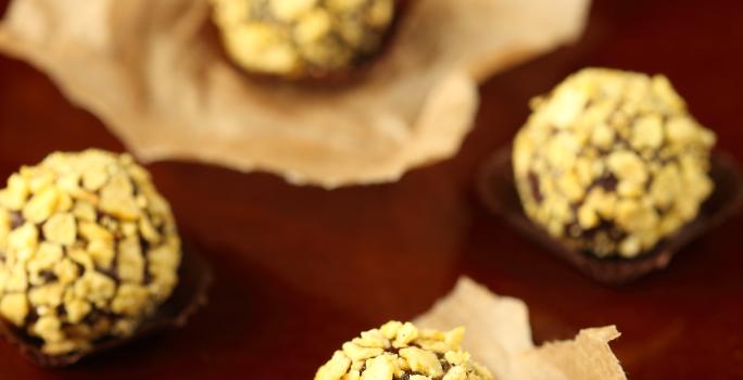 Fotografia de cinco brigadeiros enrolados e passados em nozes, alguns em forminhas, outros sobre um papel manteiga. Ao fundo, forminhas empilhadas, sobre uma mesa marrom em tom escuro.