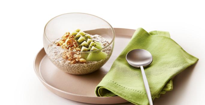 Foto da receita de Overnight Oats de Kiwi. Observa-se um prato de cerâmica marrom com um recipiente de vidro em cima com a receita. Ao lado direito, um guardanapo verde com uma colherinha.