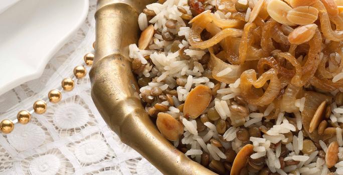 Fotografia em tons de marrom e dourado em uma mesa de madeira com uma toalha branca de renda com um recipiente redondo fundo e grande dourado com o arroz de festa com lentilhas.
