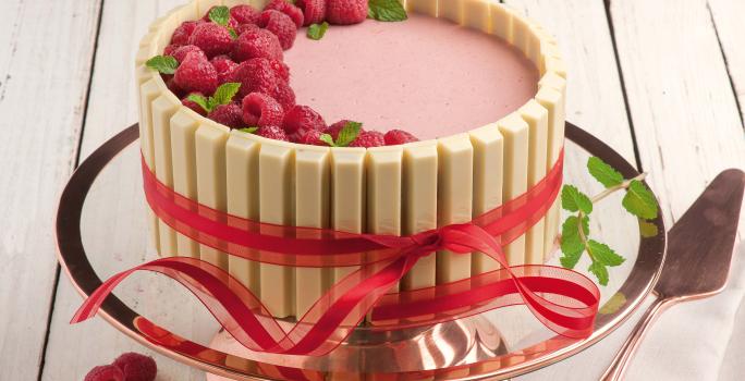 Fotografia em tons de rosa em uma bancada de madeira clara, um suporte para bolo em vidro rosado, com a mousse de framboesa com KitKat Branco ao redor, formando uma linda torta.