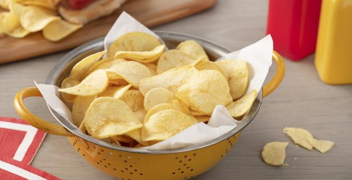 Fotografia em tons de vermelho em uma bancada de madeira clara, um pano vermelho com listra branca, uma tábua de madeira com um cachorro-quente e batatas chips ao lado. Ao centro, um recipiente redondo amarelo com muitas batatas chips.