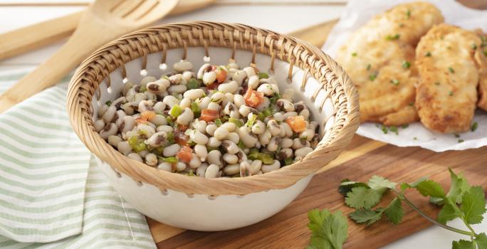 Fotografia em tons de verde em uma bancada de madeira clara, um pano verde listrado, uma tábua de madeira com um recipiente branco com detalhes beges redondo fundo com a salada do feijão-de-corda com vegetais dentro dele.