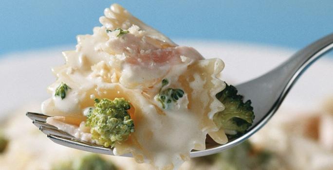 Fotografia em tons de azul, branco e verde, ao centro porção de macarrão com brócolis ao creme suspenso em um garfo, abaixo prato branco com macarrão, creme e brócolis, tudo sobre bancada azul.