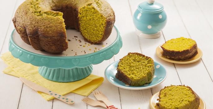 Fotografia em tons de amarelo em uma bancada de madeira de cor branca. Ao centro, um pano amarelo com uma boleira verde em cima contendo o bolo. Ao lado, há 3 pires contendo 3 fatias do bolo e ao fundo, uma chaleira.