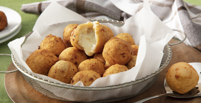 Foto da receita de bolinho de batata servida em diversos bolinho dentro de uma peneira de metal coberta com guardanapos sobre uma tábua de madeira com uma colher prateada ao lado com um bolinho. Ao fundo um paninho cinza quadriculado