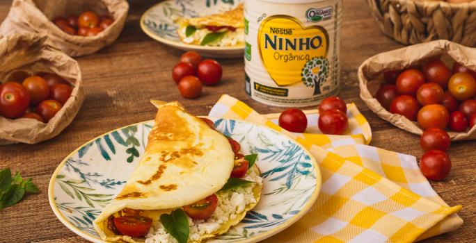 Fotografia em tons de amarelo e vermelho de uma bancada de madeira com um pano xadrez amarelo e branco, uma prato com uma crepioca recheada de queijo e tomate. Ao fundo tomates-cereja, uma lata de NINHO e ovos.